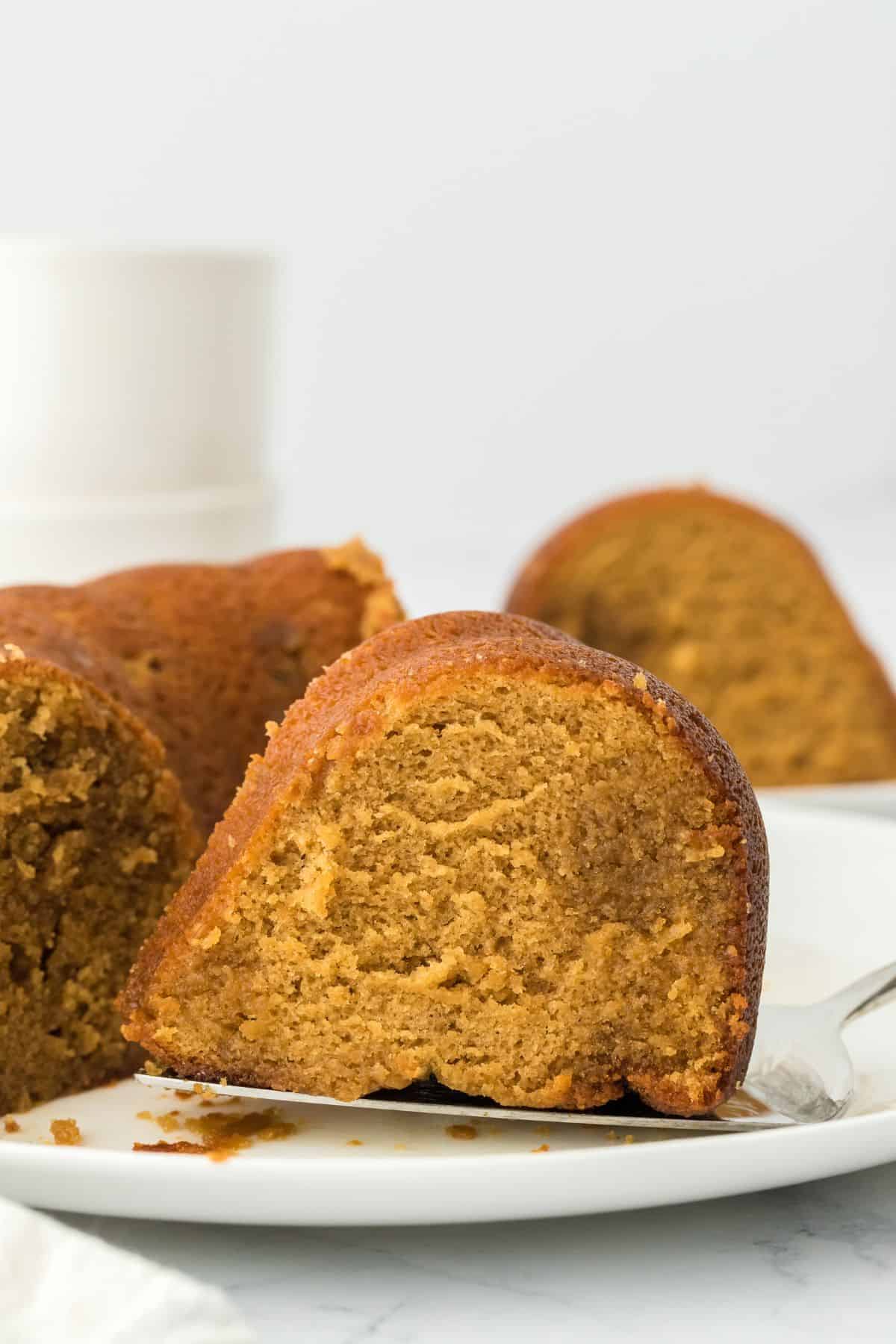 A slice of rum cake being held up on a cake server, with the rest of the cake visible in the background